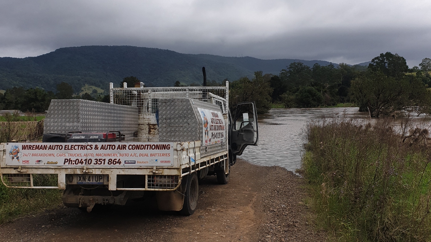 The Wireman work ute by a flooded river, its tray sign reading 24/7 breakdown, cars, trucks, auto machinery, auto A/C, dual batteries