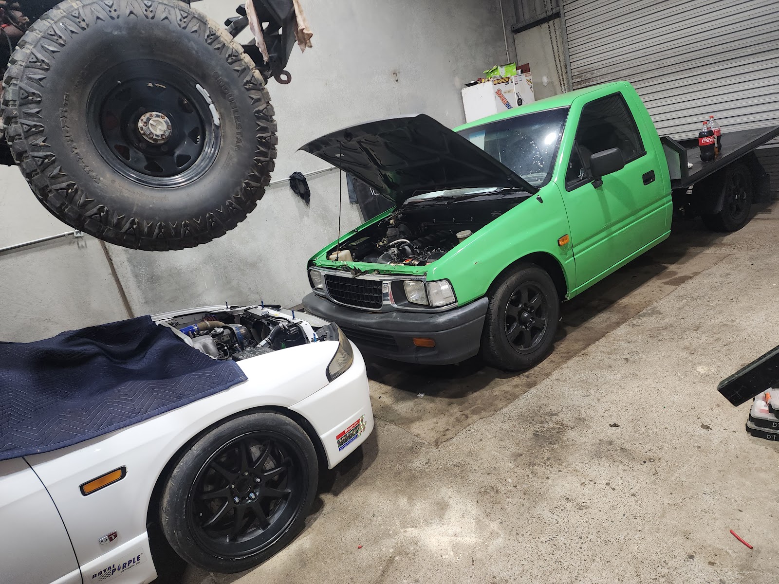 Green ute and white car inside the Wireman shed with a spare wheel hanging from the roof